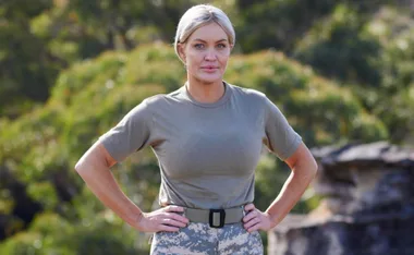 Person in a military-style outfit stands confidently outdoors with hands on hips, set against a blurred green background.