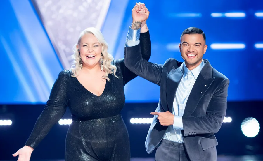 A smiling woman in a black dress and a man in a suit celebrate with raised arms on stage amidst vibrant blue lighting.