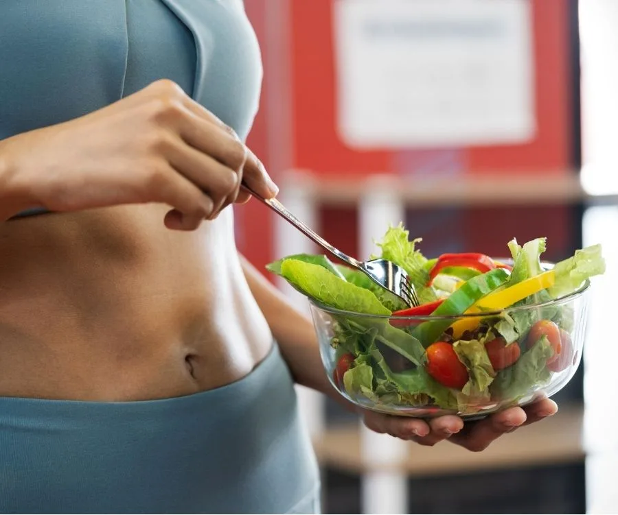 Person in workout attire holding and eating a fresh salad with mixed vegetables from a glass bowl.