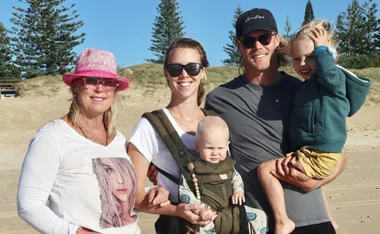 Family posing on a sunny beach, wearing casual clothes and sunglasses, with pine trees in the background.