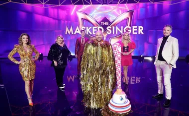 The Masked Singer stage with hosts and a contestant in a gold fringed costume, holding a novelty plunger.