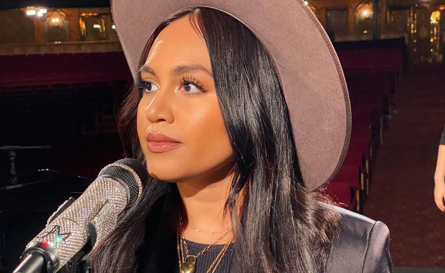 Singer wearing a wide-brimmed hat, performing on stage with a microphone in a dimly lit venue.