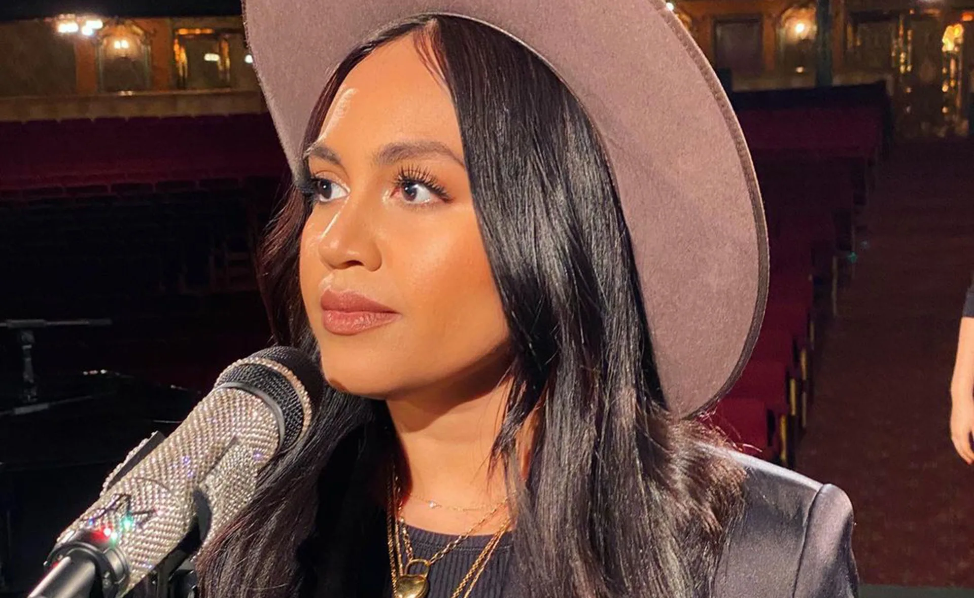 Singer wearing a wide-brimmed hat, performing on stage with a microphone in a dimly lit venue.