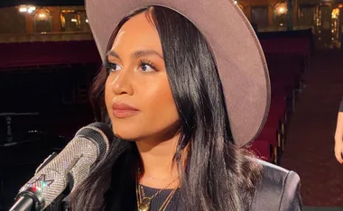 Singer wearing a wide-brimmed hat, performing on stage with a microphone in a dimly lit venue.