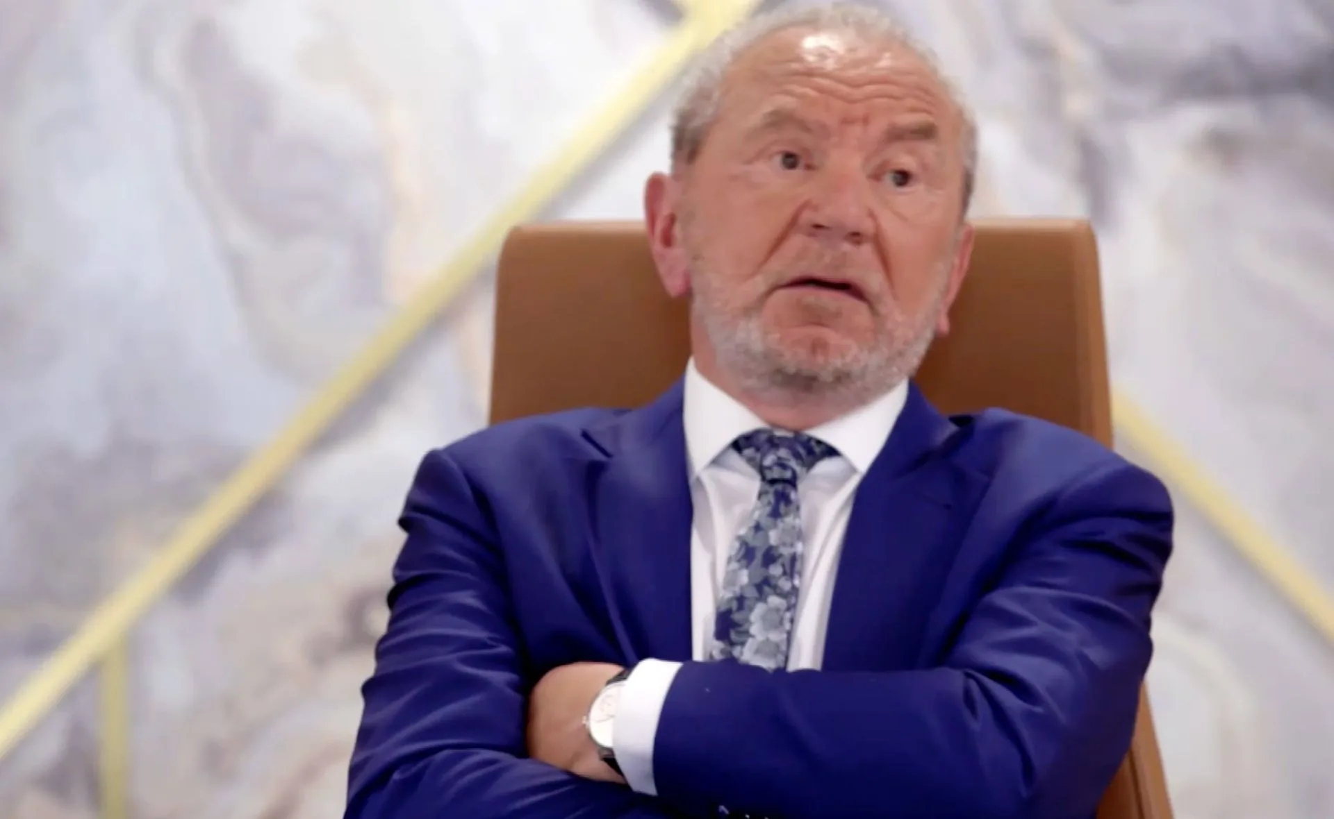 An older man in a sharp blue suit and patterned tie sits with arms crossed, looking pensive.