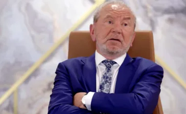 An older man in a sharp blue suit and patterned tie sits with arms crossed, looking pensive.