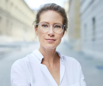 Middle-aged woman with glasses, standing on a street, looking confident.