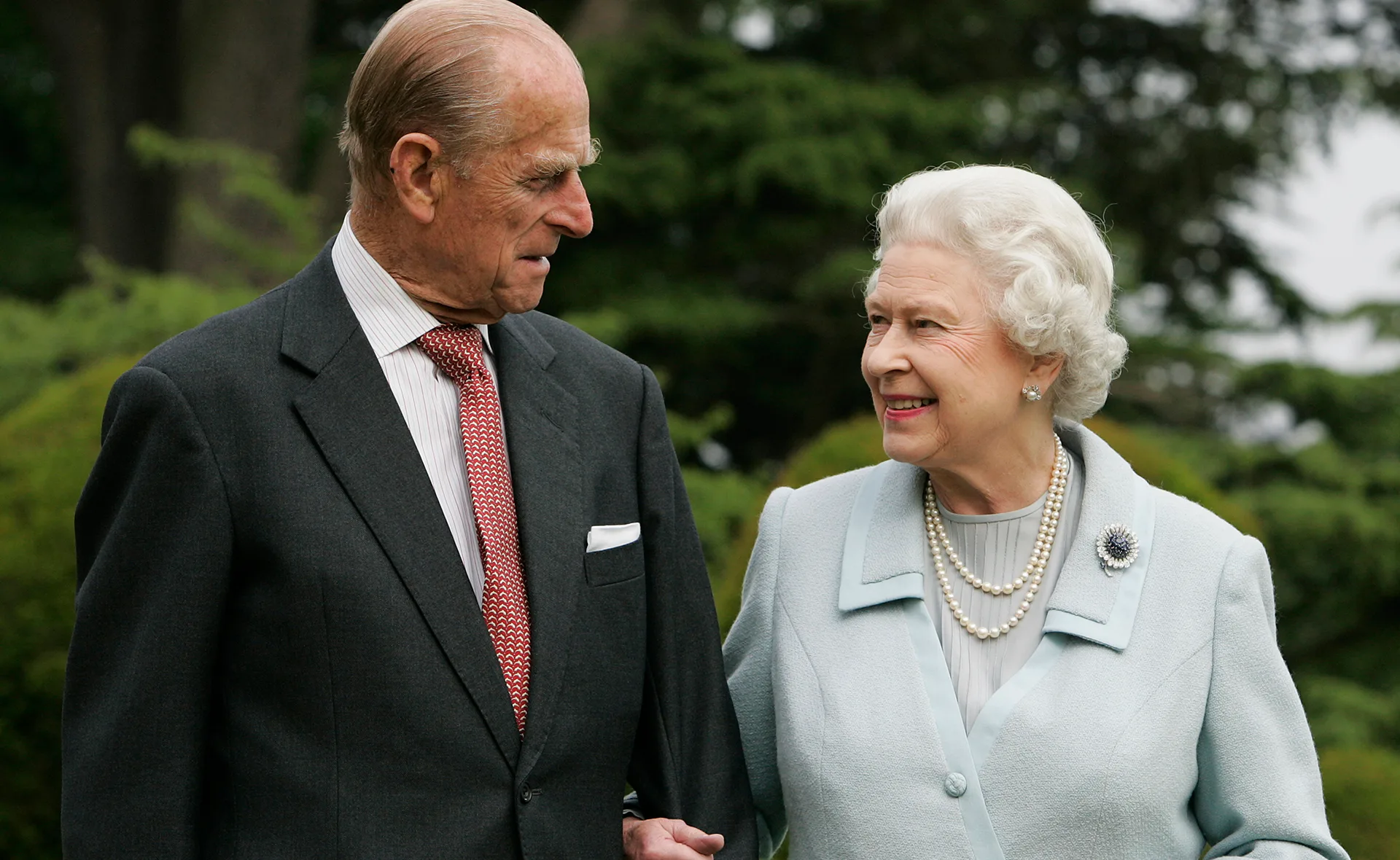 Elderly couple walking together in a garden, smiling and holding hands, with lush greenery in the background.