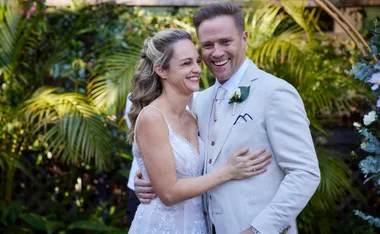 Bride and groom in elegant attire embrace and smile outdoors, surrounded by greenery.