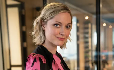 A woman with short blonde hair smiles slightly, wearing a pink and black patterned top, inside an office setting.