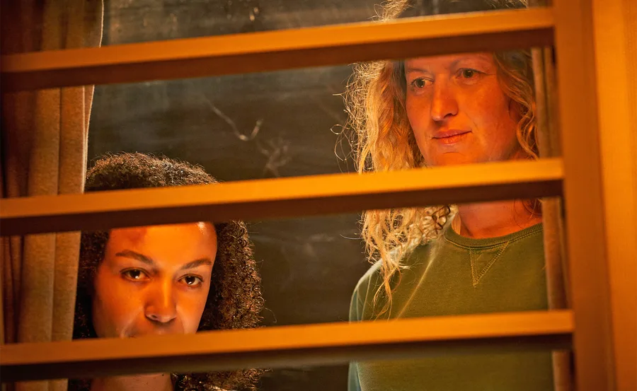 Two individuals look through slatted blinds with focused expressions in a dimly lit setting.