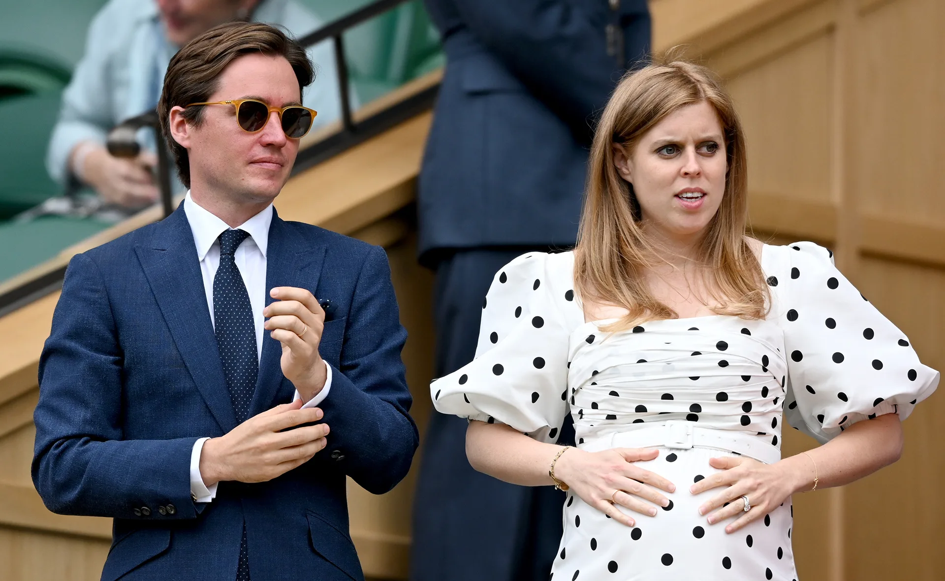 A woman in a polka dot dress holds her baby bump while a man in a suit stands beside her at an outdoor event.