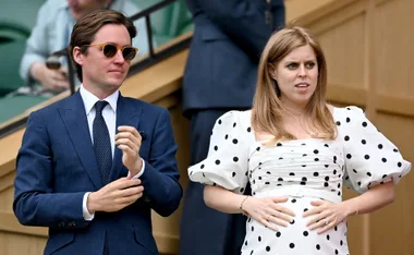 A woman in a polka dot dress holds her baby bump while a man in a suit stands beside her at an outdoor event.
