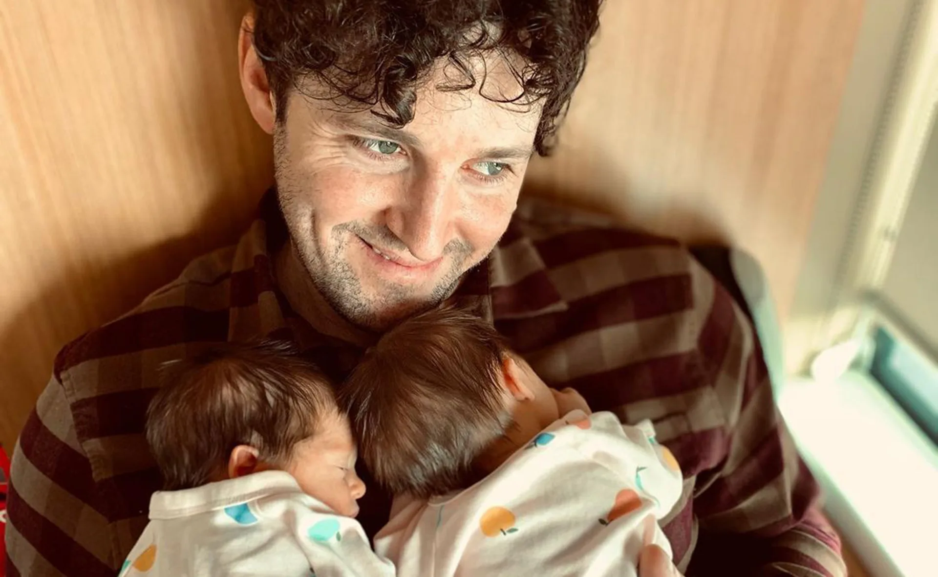 Father smiling while holding two newborn babies wrapped in patterned blankets.