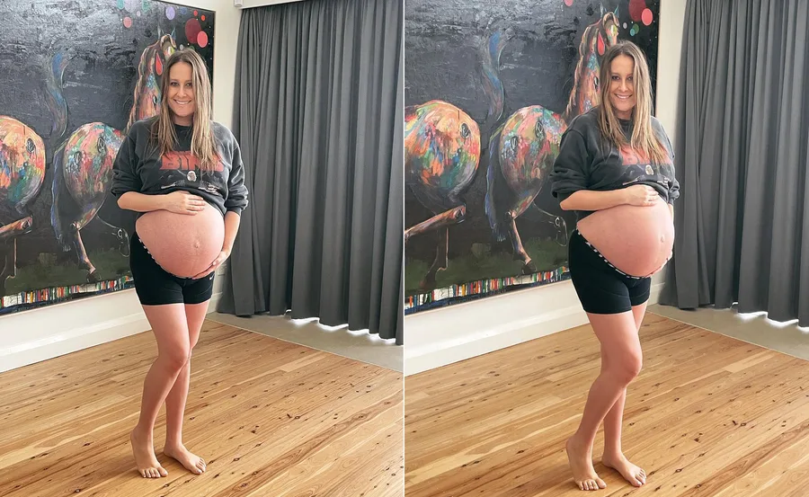 A pregnant woman smiling, holding her belly, standing barefoot in front of a colorful art piece.