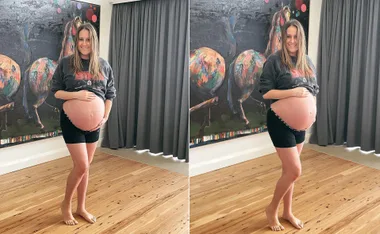 A pregnant woman smiling, holding her belly, standing barefoot in front of a colorful art piece.