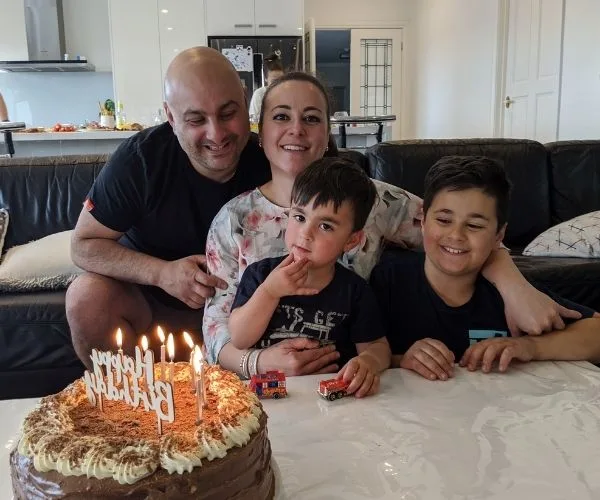 Family celebrating birthday with chocolate cake, lit candles; parents and two children smiling at the table.