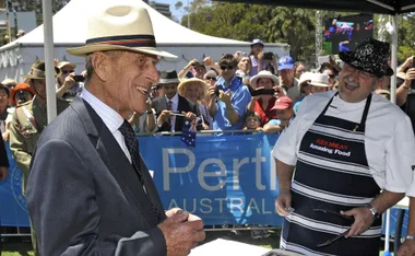 Prince Philip laughing with a chef at an outdoor event in Perth, Australia, surrounded by a crowd with cameras.