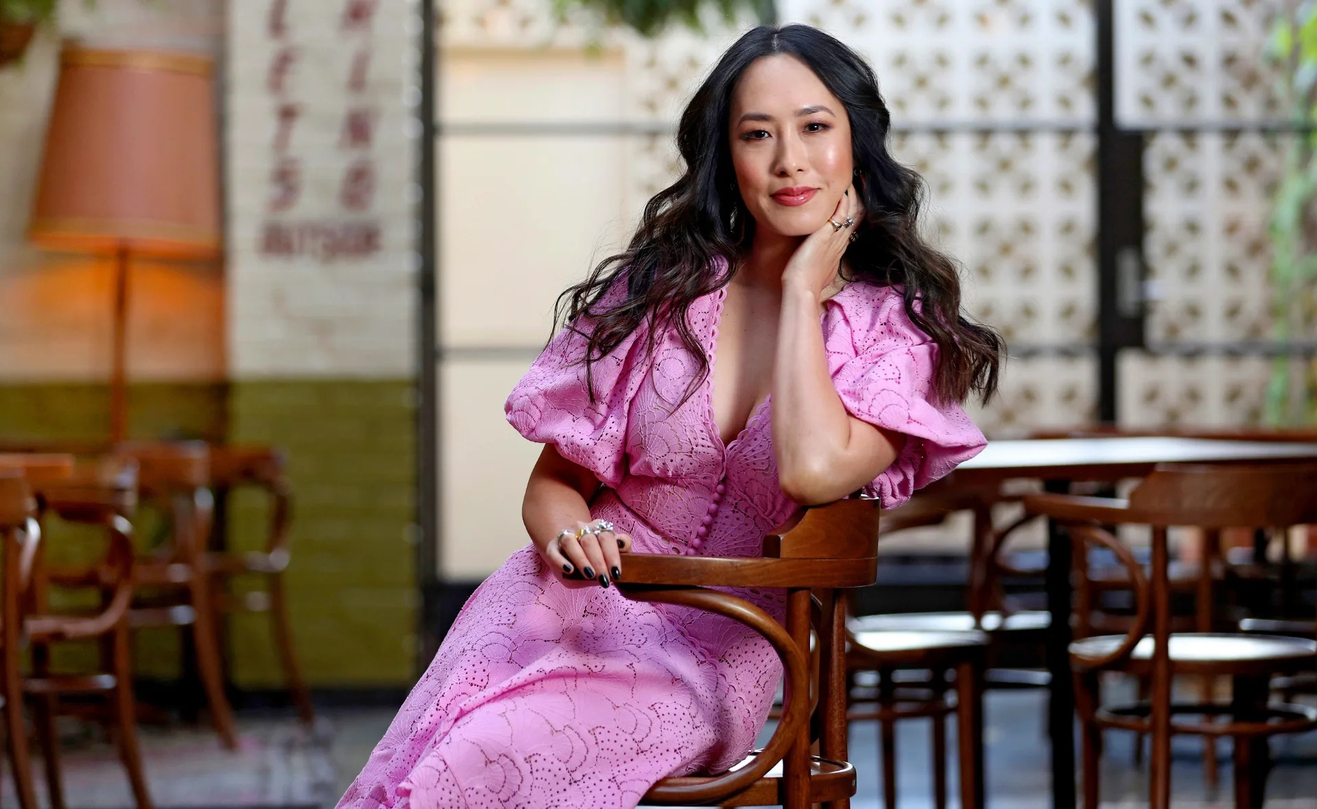 A woman in a pink dress sits on a wooden chair, posing confidently with her hand on her face in a stylish setting.