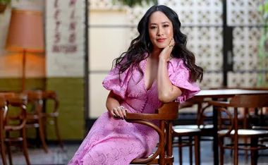 A woman in a pink dress sits on a wooden chair, posing confidently with her hand on her face in a stylish setting.