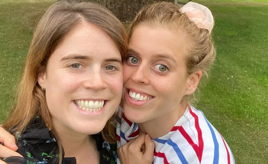 Two women smiling closely together, both with light hair, outdoors with green grass in the background.