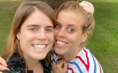 Two women smiling closely together, both with light hair, outdoors with green grass in the background.