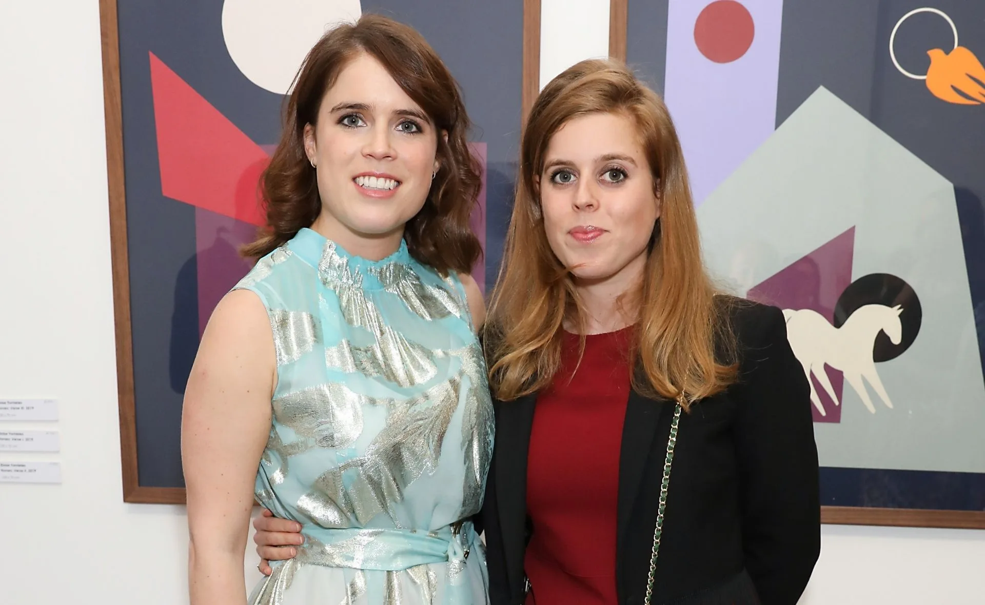 Two women smiling, one in a light blue dress and the other in a red dress with a black jacket, stand in front of artwork.
