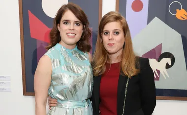 Two women smiling, one in a light blue dress and the other in a red dress with a black jacket, stand in front of artwork.