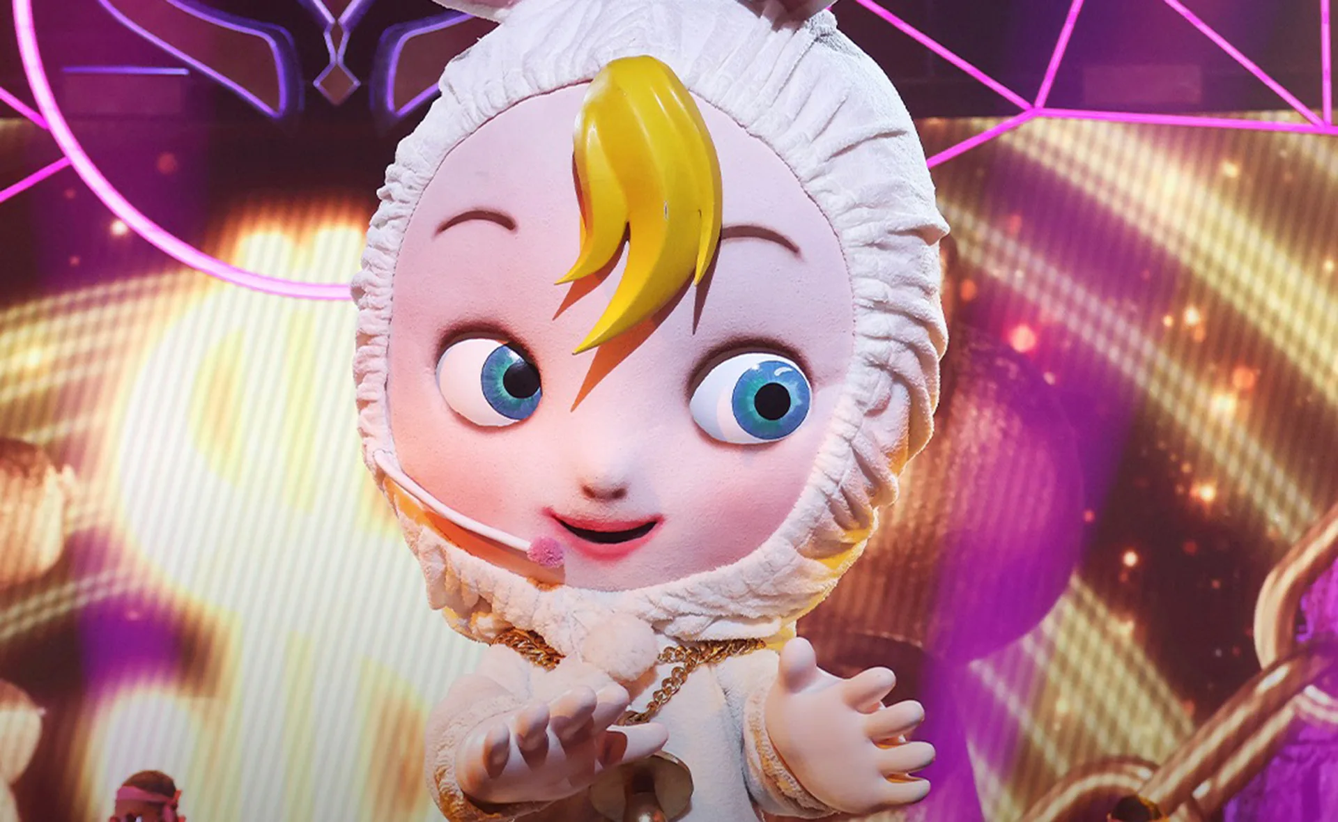 Cartoonish baby character with blue eyes and yellow hair on a stage with colorful lights in the background.