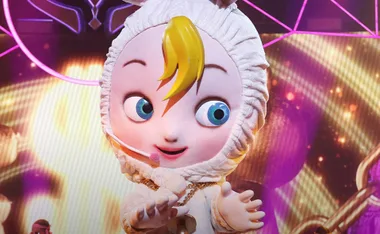 Cartoonish baby character with blue eyes and yellow hair on a stage with colorful lights in the background.