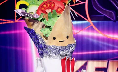 Kebab costume with smiling face, wrapped in foil, colorful vegetables, on stage with vibrant pink and purple background.