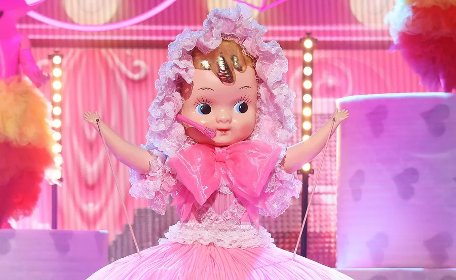 A person in a pink baby doll costume with large eyes and a big bow, standing on a colorful stage.
