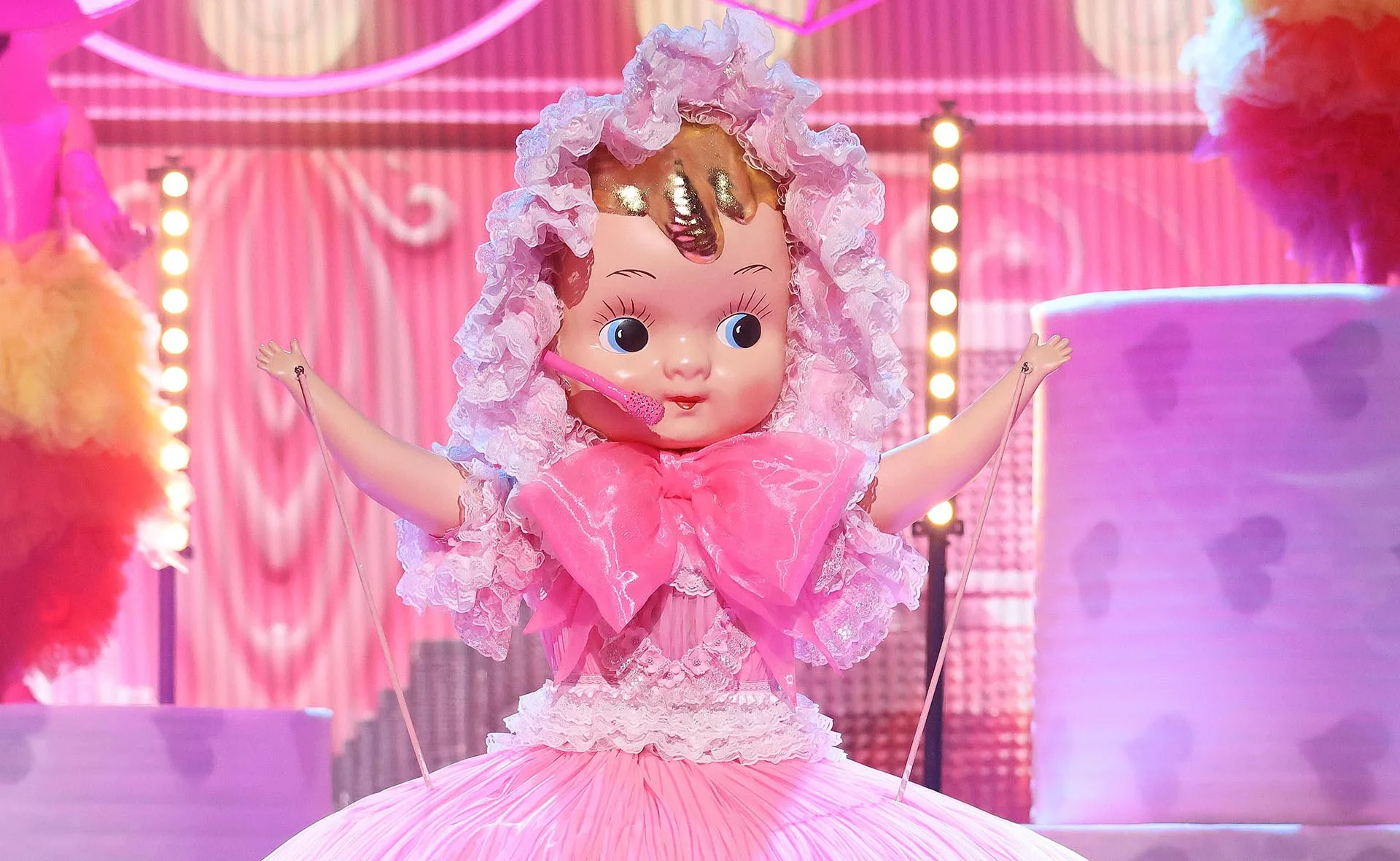 A person in a pink baby doll costume with large eyes and a big bow, standing on a colorful stage.