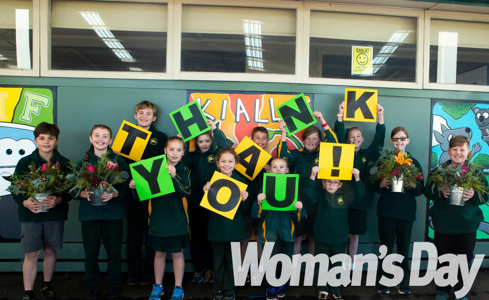 Children holding "THANK YOU!" signs and flowers, smiling at the camera in front of a green wall, "Woman's Day" logo visible.
