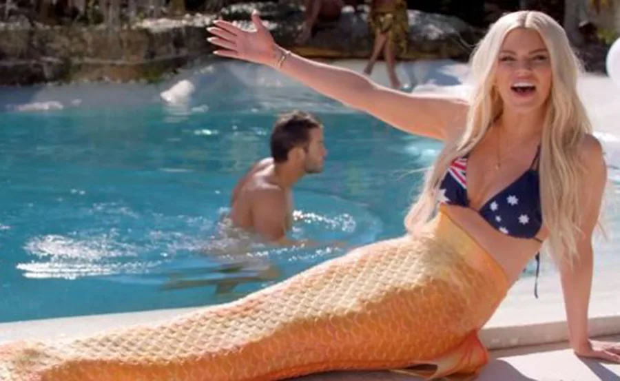 Woman in a mermaid tail and flag bikini near a pool, smiling and raising an arm; background includes a man in water.