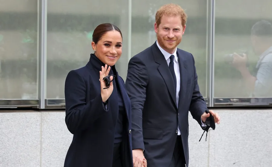 A couple in formal attire smiles and holds hands, with the woman waving.