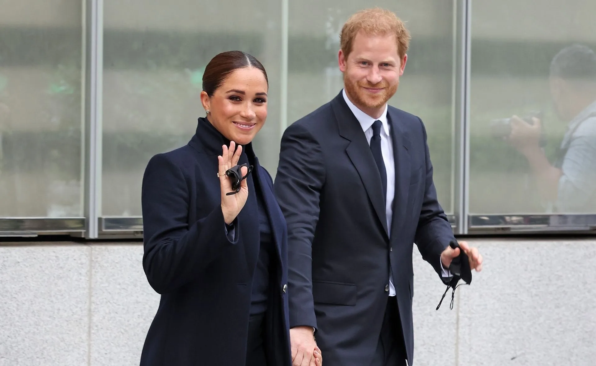 A couple in formal attire smiles and holds hands, with the woman waving.