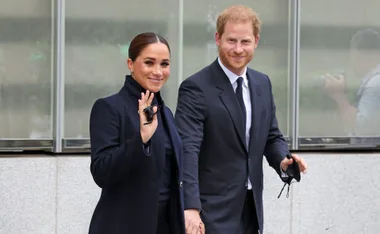 A couple in formal attire smiles and holds hands, with the woman waving.
