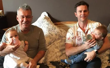 Two men, each holding a baby with bottles, sitting on a couch with decorative pillows in the background.