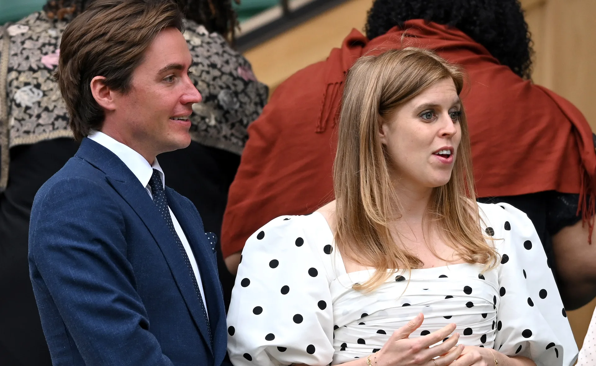 Man in a blue suit with a polka dot tie, standing next to a woman in a white dress with black polka dots, talking.