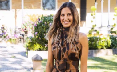 A woman in a sleeveless, patterned dress stands outside in a garden with flowers and shrubs in the background.