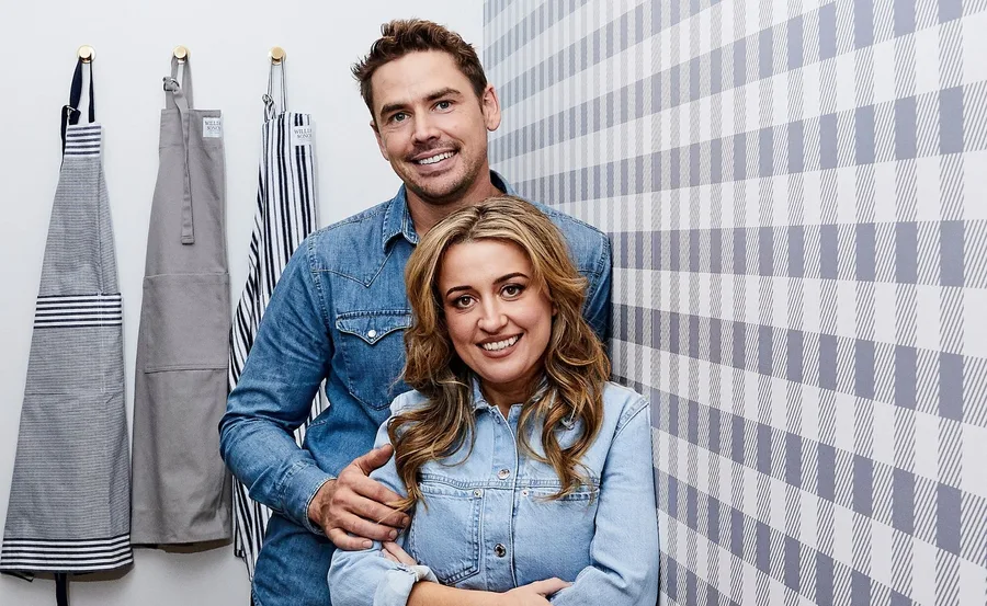 A smiling couple in denim shirts poses against a checkered wall beside hanging aprons.