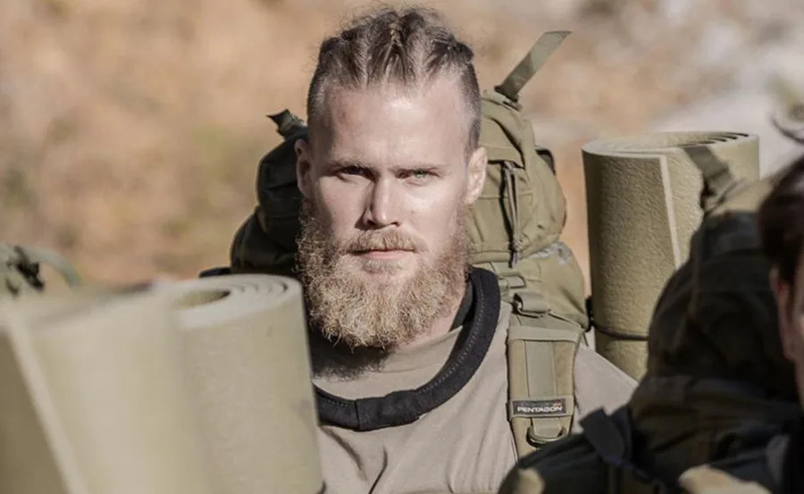 Man with braided hair and a beard, wearing military gear with backpacks and rolled-up mats, in an outdoor setting.