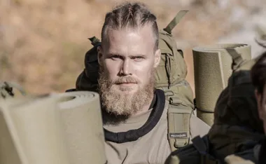 Man with braided hair and a beard, wearing military gear with backpacks and rolled-up mats, in an outdoor setting.
