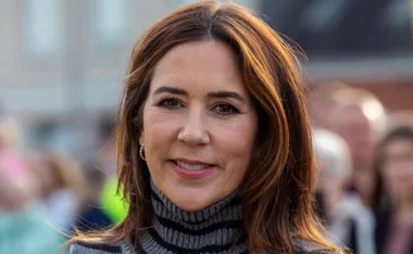A woman with long brown hair smiles, wearing a striped turtleneck sweater, with a blurred crowd in the background.