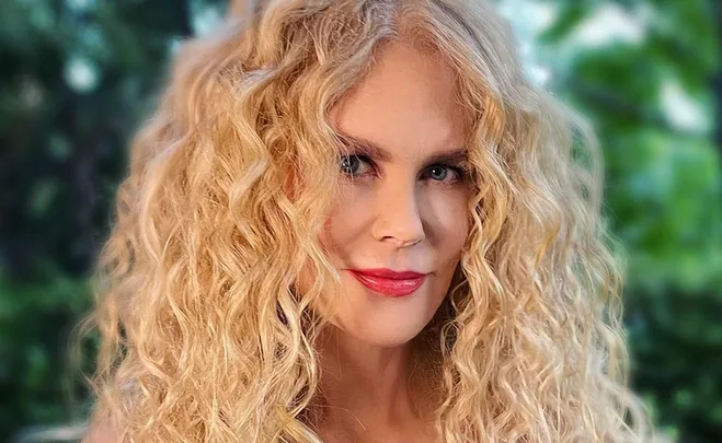 A person with long, curly blonde hair and bright red lipstick, smiling slightly against a blurred green background.