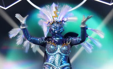 A performer in an elaborate blue and silver costume with glitter and a lightning-themed headdress on a vibrant stage.
