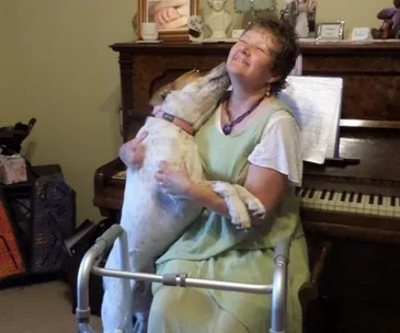 Woman in green dress sitting by a piano, hugging a large dog with a walker in front of her, smiling joyfully.