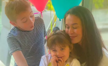 A woman embraces a smiling young girl, with a boy near them and colorful balloons in the background.