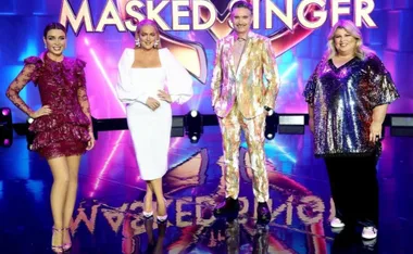 Four judges in colorful outfits stand on "Masked Singer" stage with bright lights and show logo in background.